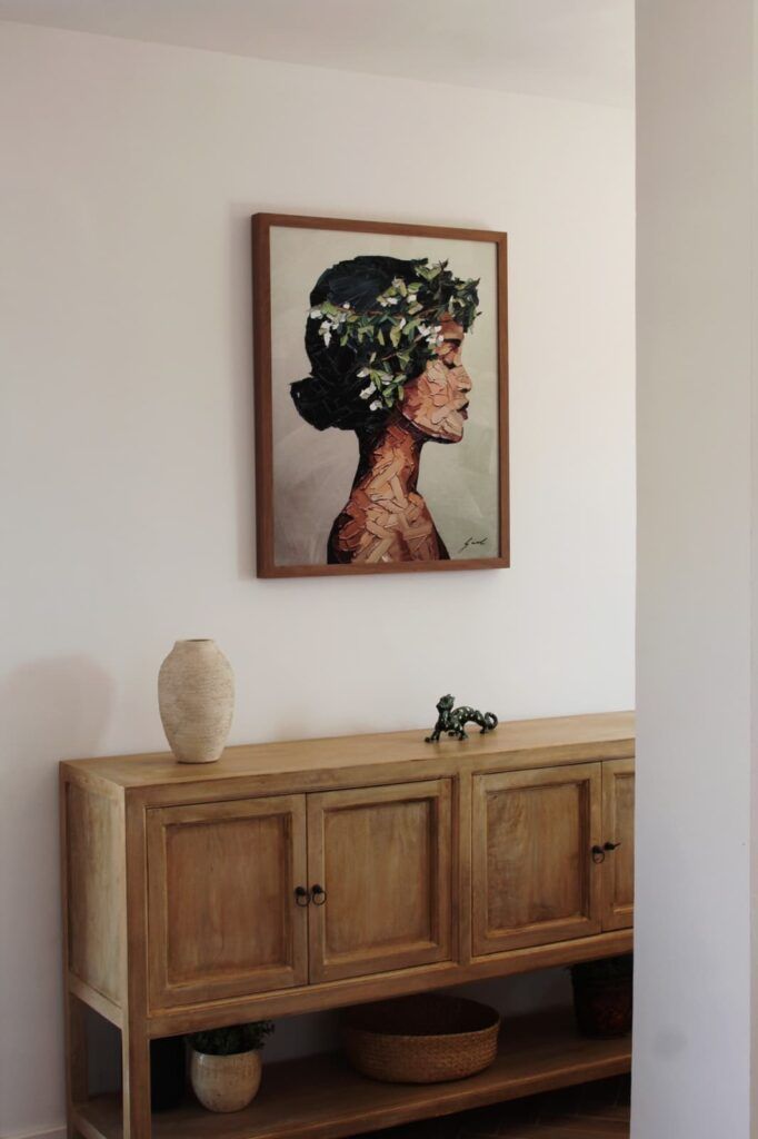 Un aparador de madera con un jarrón, una pequeña escultura y una cesta se apoya en una pared blanca. Encima cuelga un cuadro enmarcado de una mujer de perfil con el pelo corto y oscuro y hojas verdes en el cabello.