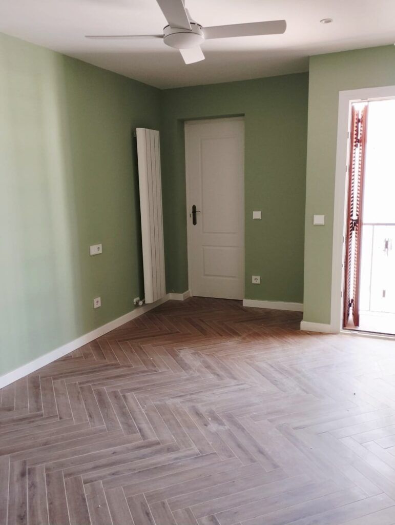 Habitación vacía con paredes verde pálido, ventilador de techo blanco, puerta blanca, radiador vertical alto y suelo de madera con dibujo de espiga; la luz del sol entra por una ventana con contraventanas abiertas a la derecha.