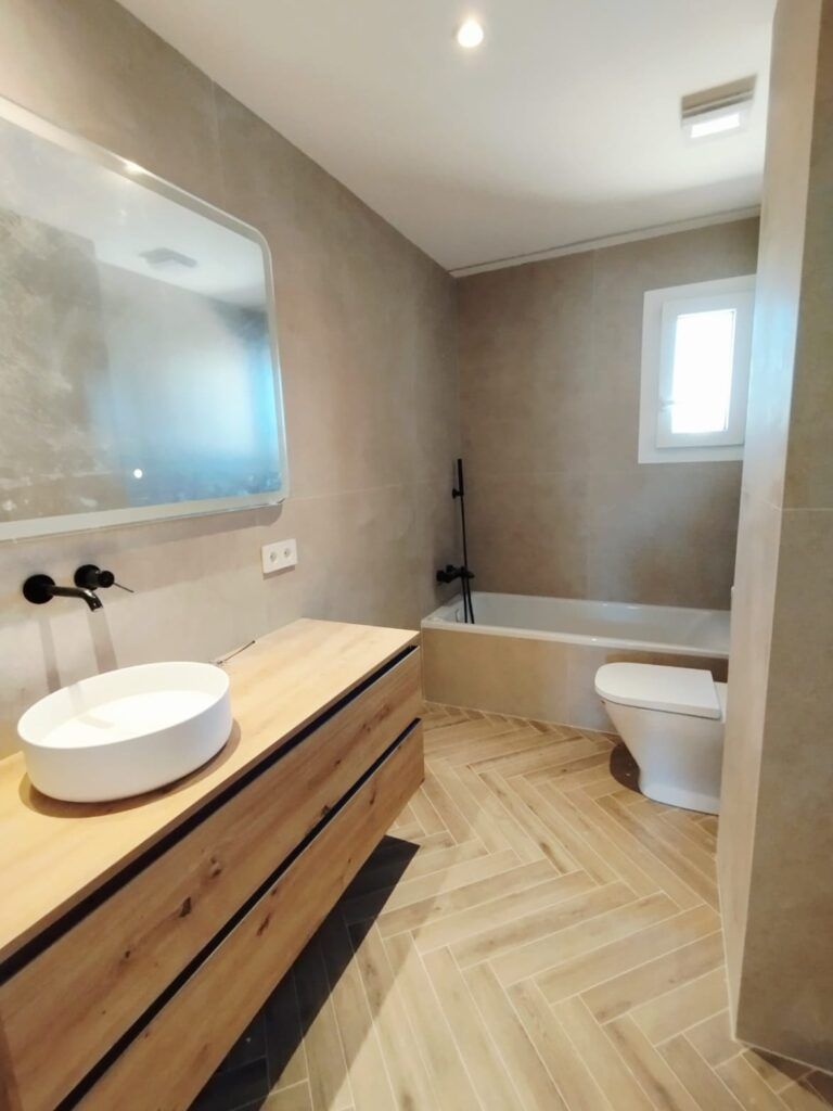 Moderno cuarto de baño con paredes de color beige, un mueble de madera con lavabo redondo blanco y grifo negro, un gran espejo, un inodoro, una bañera con ducha negra y una pequeña ventana por la que entra luz natural.
