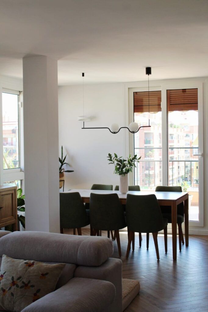 Un comedor moderno con una mesa de madera, seis sillas verdes, un centro de mesa con plantas y una lámpara minimalista, junto a grandes ventanales con vistas a la ciudad. En primer plano, un sofá gris con cojín.