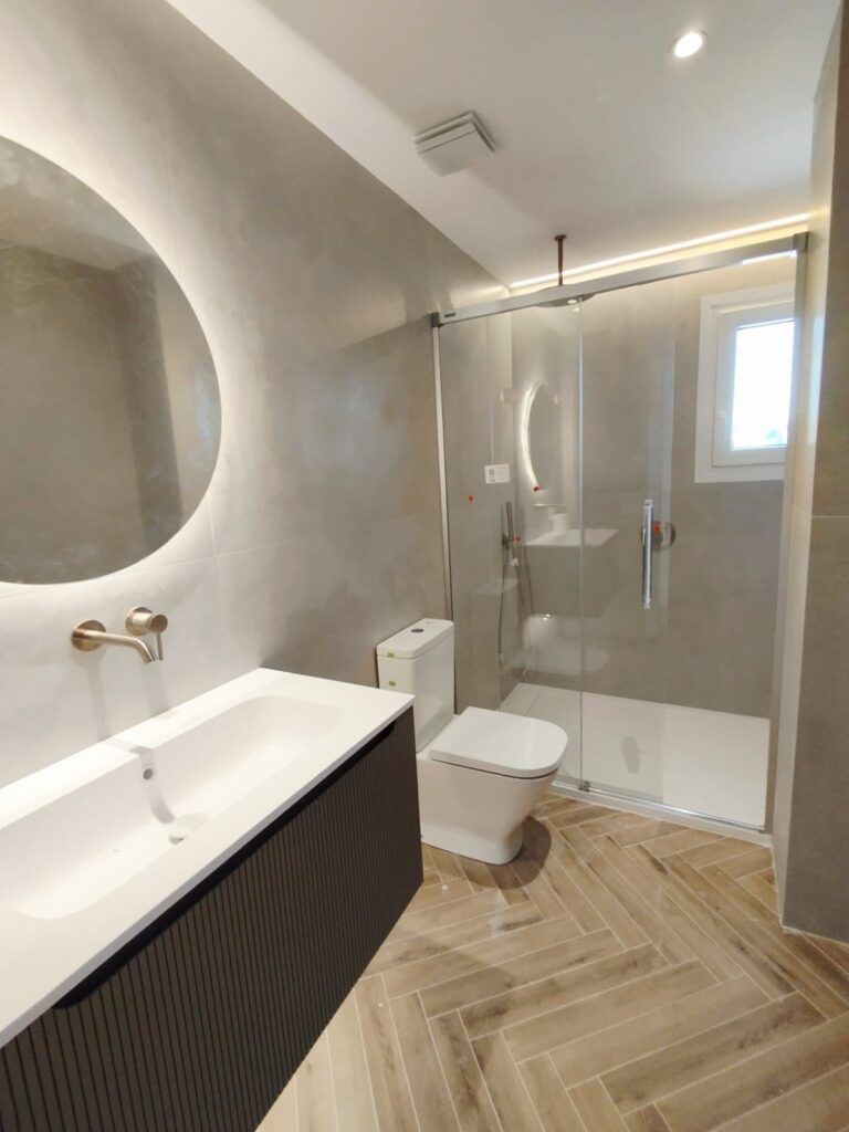 Moderno cuarto de baño con lavabo flotante, espejo redondo, grifo montado en la pared, inodoro, mampara de ducha de cristal y baldosas de madera con dibujo de espiga. Las paredes gris claro y una pequeña ventana añaden luminosidad.