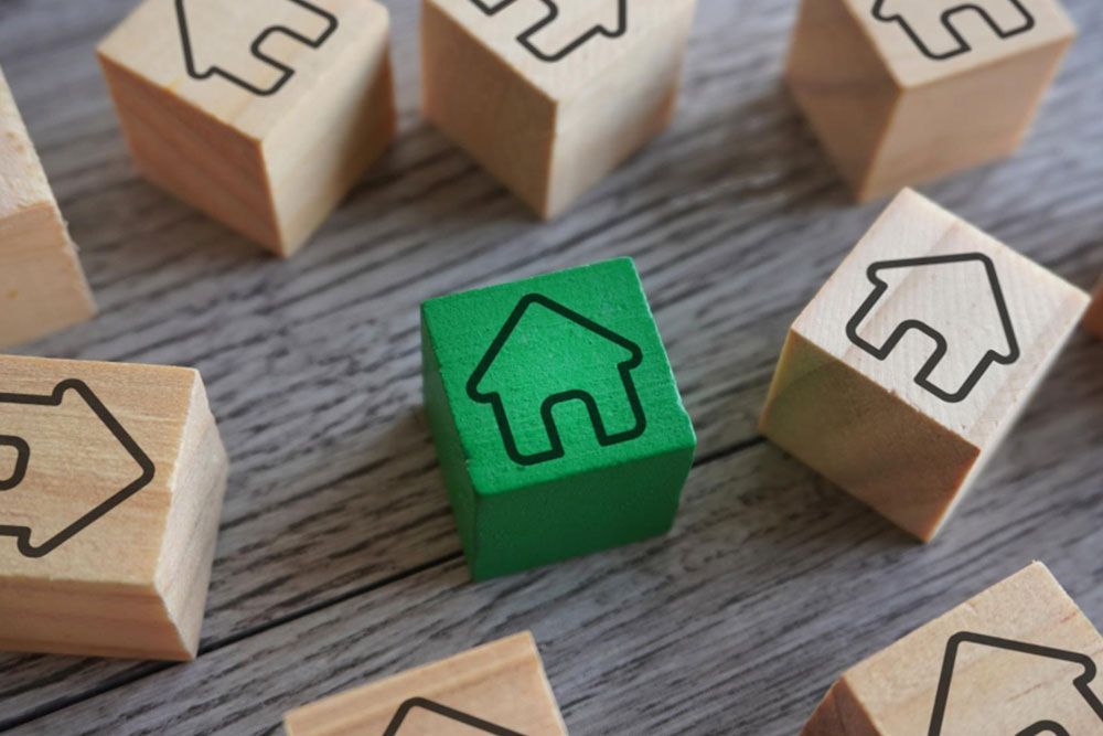 Un grupo de bloques de madera con iconos de casas, un bloque en el centro es verde mientras que los otros son de color madera natural, todos dispuestos sobre una superficie de madera gris.