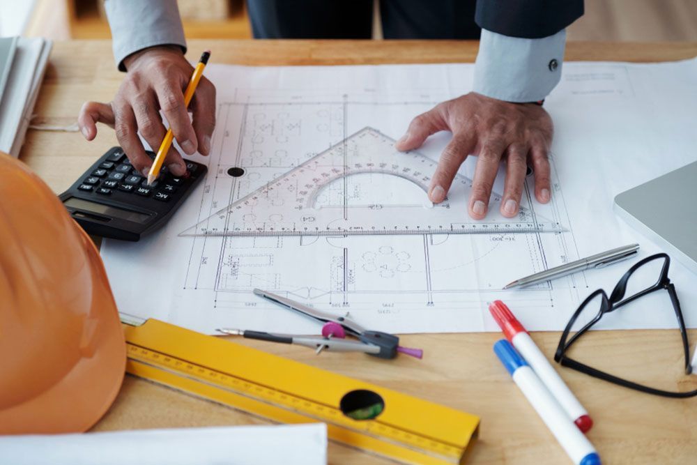 Las manos de un ingeniero trabajan en planos con un lápiz, una regla y una calculadora sobre un escritorio. Alrededor del espacio de trabajo también se ven un casco naranja, gafas, un ordenador portátil, un nivel de burbuja y rotuladores.