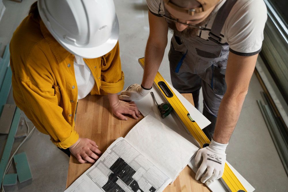 Dos personas con ropa de trabajo y cascos examinan un plano de construcción sobre una mesa de madera. Cerca hay una cinta métrica y un nivel de burbuja, lo que indica que se está realizando un proyecto de planificación o construcción.