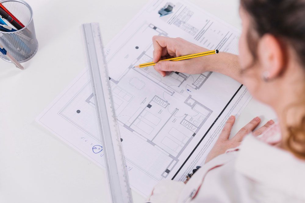 Una persona dibuja en un plano arquitectónico con un lápiz, utilizando una regla para mayor precisión. En el escritorio, junto al dibujo, se ve una taza con bolígrafos y lápices.
