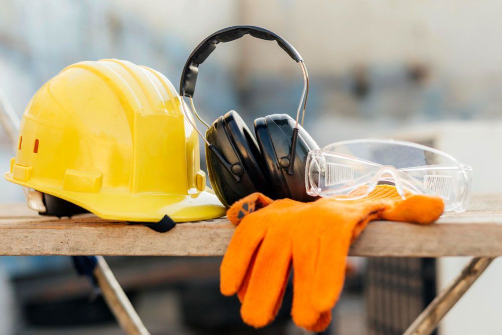 Un casco amarillo, unas orejeras negras, unas gafas de seguridad transparentes y unos guantes de trabajo naranjas están ordenados sobre una superficie de madera, representando el equipo de seguridad esencial para la construcción.